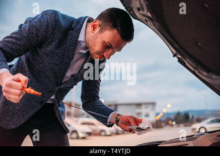 Young manager formally dressed checking his car oil at sunset while leaning under the car's hood. Stock Photo
