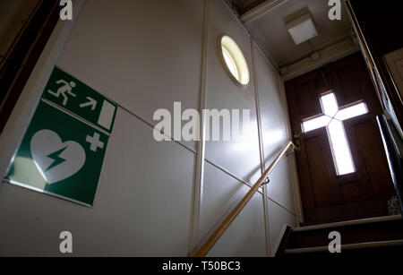 Hamburg, Germany. 14th Apr, 2019. Light shines through a glass cross in the entrance door of the River Ship's Church. On the wall hang pictograms for the emergency exit and rescue equipment for heart problems. Germany's only floating church has been anchored in Hamburg since 1952. The Evangelical Lutheran Church once had the boat converted into a floating church. Credit: Christian Charisius/dpa/Alamy Live News Stock Photo