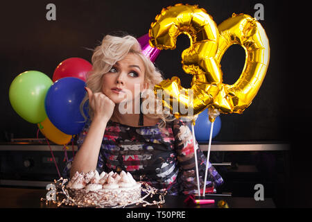 Anniversary birthday. Young beautiful blonde sitting in front of a birthday cake and sad, young woman celebrates her birthday alone. Stock Photo