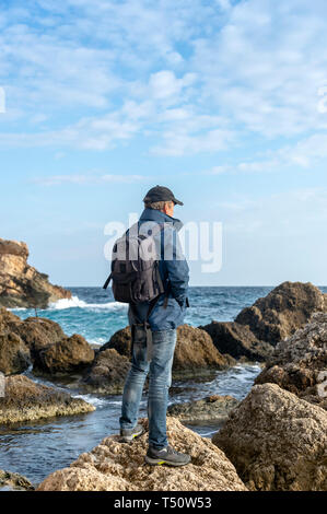 man wearing a backpack standing on a rock looking out to sea Stock Photo