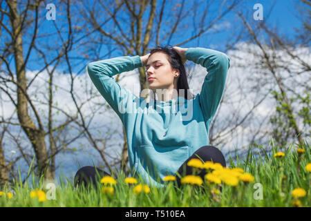 Girl in hoodie sitting on green meadow among flowers and meditating in nature background. Woman practicing meditation. Eyes closed. Stock Photo