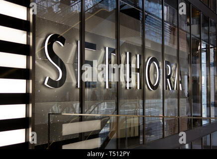 Illuminated Sephora sign on the side of the French multinational personal care and beauty chain's store at the mall in the new Hudson Yards complex, opened earlier that month; the paneled glass in front reflecting people and escalators, the company's signature black and white horizontal stripe branding to the left. Hudson Yards, New York's newest neighborhood, debuted 15 March 2019, to controversy about whether it was too elitist. The development encompasses retail, residential, restaurants, cultural institutions, public space, and the world's first Equinox hotel. Stock Photo