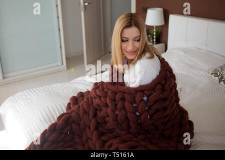An attractive young woman is wrapped in a soft fluffy blanket. Girl Sitting comfortably on the bed at home. Stock Photo