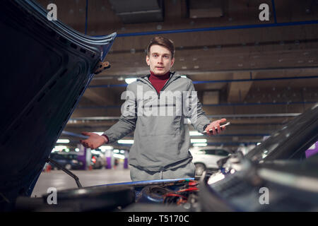 Upset man with phone in hand near broken car with open hood in underground parking Stock Photo