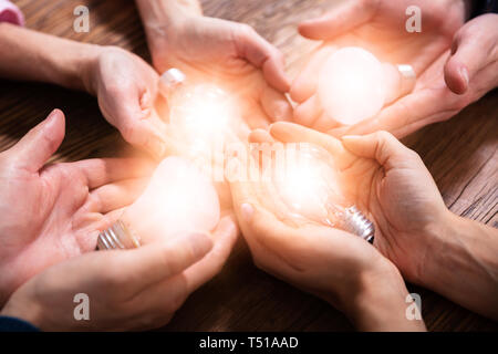 An Elevated View Of Businesspeople's Hand Holding Illuminated Light Bulbs In Hands Over Wooden Desk Stock Photo