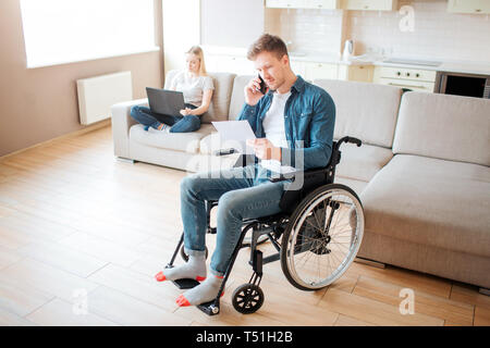 Young worker with disability in room. Holding piece of paper and talking on phone. Young woman sit behind on couch with laptop. Daylight. Couple Stock Photo