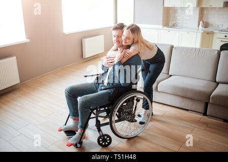 Cheerful emotions. Young man with disability sitting on wheelchair. Woman stand behind and embrace him. Lovely couple spend time together Stock Photo