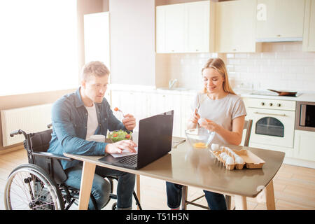 Concentrated young man on wheelchair working with laptop and eating salad. Studying with disability and inclusiveness. Guy with special needs. Young w Stock Photo