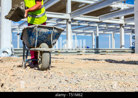 Construction worker is using shovel to unloading cement from wheelbarrow on construction site. Stock Photo
