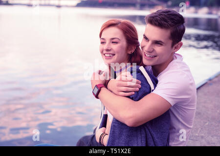 Strong smiling guy tightly hugging his pretty ginger girlfriend Stock Photo