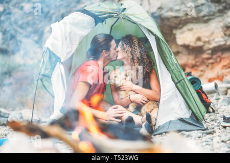 Travel couple kissing while sitting in the tent with their pet - Happy man and woman having a romantic moment vacation camping around rock mountain Stock Photo