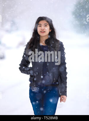 Biracial teen girl wearing black leather jacket and blue jeans walking down middle of street in heavy snowstorm