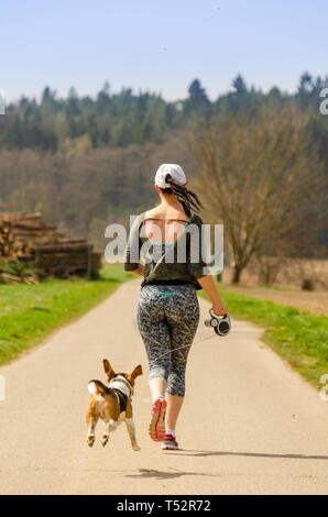 Girl running with dog outdoors in nature on a road to forest. Sunny day countryside sunset. Stock Photo