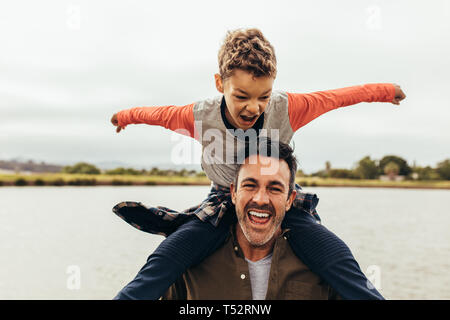 Boy sitting on the shoulders of his father and playing. Father and son having fun spending time together near a lake. Stock Photo