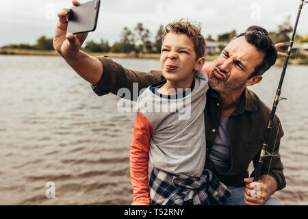 Close up of father and son taking a selfie while fishing near a lake. Happy father and son making faces while takes a selfie near the lake holding a f Stock Photo