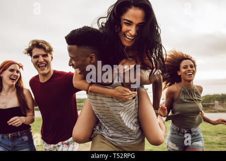 Smiling group of friends enjoying on weekend. Multi-ethnic men and women having fun outdoors. Man carrying woman in his back and running with friends 