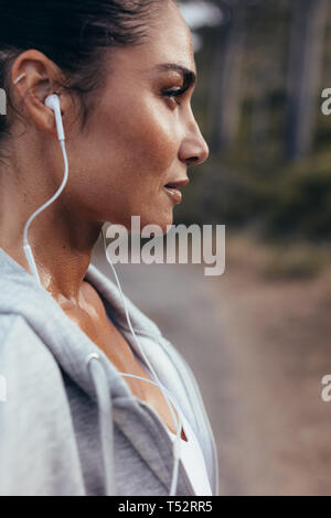 Close up of a female runner with earphones standing outdoors. Woman listening to music during workout outdoors in morning. Stock Photo
