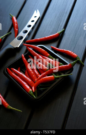 Red chilli pods and a vegetable and cheese knife lie on a black wooden surface. Daylight. Vertical image. Stock Photo