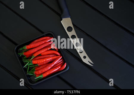 A knife for cheese and vegetables next to a box of pods of red chilli pepper lie on a black wooden surface. Daylight Stock Photo