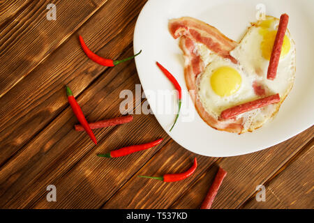Omelet with bacon, small sausages and red chili peppers in a white plate on a natural wooden surface made of pine boards. Daylight. View from above. Stock Photo