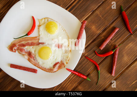 Omelet with bacon, red chilli peppers and small sausages in a white plate on a natural wooden surface of pine boards. Daylight. View from above. Stock Photo