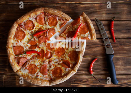 A cheese knife and homemade pizza with pepperoni and red chili peppers lie on a natural wooden surface made of pine boards. Daylight. Cut off a piece. Stock Photo