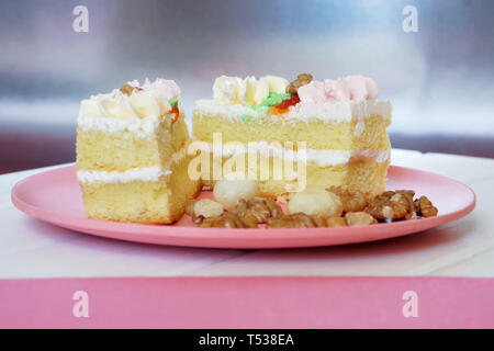 Split biscuit cake with cream wraps, macadamia nuts and walnuts in a plate on a white and pink background. Daylight. Stock Photo