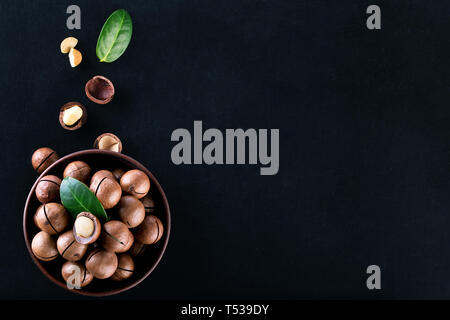 Macadamia nuts in a clay plate with grains and leaves on a black background. View from above Stock Photo