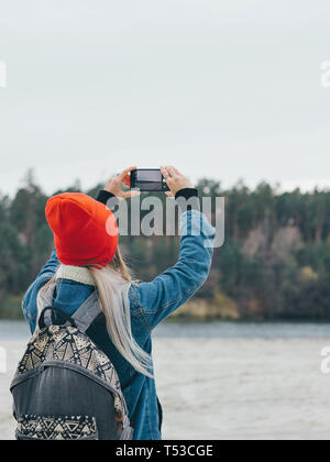 Walk along the river A girl in a red cap with a backpack is taking pictures of the riverside on a smartphone Stock Photo