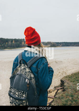 Walk along the river A girl in a red cap and with a backpack on her back is walking along the hilly bank of the river in autumn Stock Photo