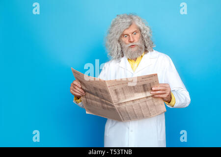 Old long-haired man wearing doctor's gown studio isolated on blue wall standing holding newspaper reading news shocked Stock Photo