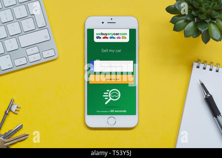An iPhone showing the We Buy Any Car website rests on a yellow background table with a keyboard, keys, notepad and plant (Editorial use only). Stock Photo