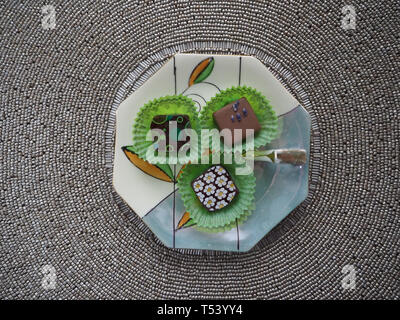 Three fancy chocolate candies in green wrappers on a porcelain dish with a beaded placemat in the background. Stock Photo