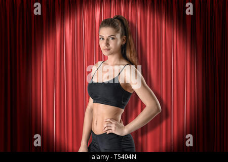 Close-up of young woman in black sport crop top and shorts standing in half-turn with one hand on hip and looking at camera against red curtain. Stock Photo