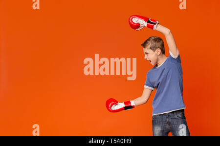 Aggressive boy punching in boxing gloves and screaming aside at empty space, orange studio background Stock Photo