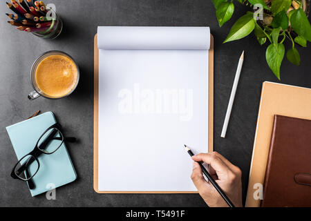 Design concept - top view of man hand hold pencil on white A4 flipped paper with brown clipboard, glasses, potted plant, coffee, pencil on black backg Stock Photo
