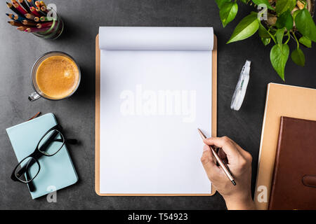 Design concept - top view of man hand hold pencil on white A4 flipped paper with brown clipboard, glasses, potted plant, coffee, pencil on black backg Stock Photo