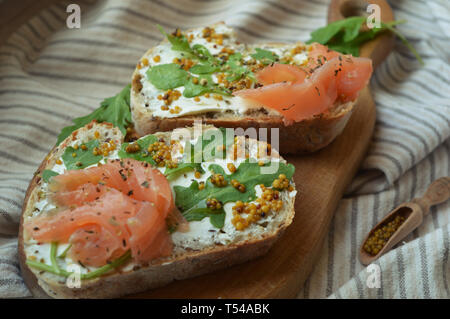 Homemade salmon sandwiches with cream cheese, mustard seeds, olive oil, arugula on toasted rye bread, served on the wooden board. Healthy trendy food. Stock Photo