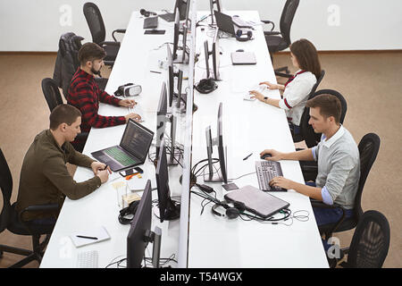 Young professionals working in modern office. Group of developers or programmers sitting at desks focused on desktops and laptops in IT company open s Stock Photo