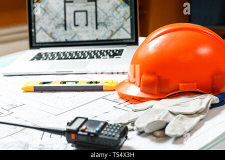 Orange hard hat, laptop  with drawings, level meter, walkie talkie and gloves lying on a  blueprints on a table. Architectural and construction concep Stock Photo