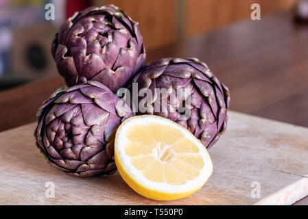 three Tuscan artichokes and a slice of lemon lit by sunlight on a wooden chopping board, dark wooden table background Stock Photo