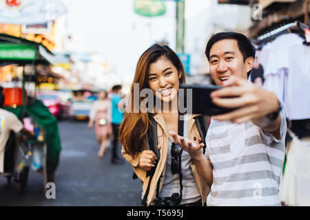 Modern young Asian man and woman exploring city while traveling and taking selfie on urban background, Bangkok, Thailand Stock Photo