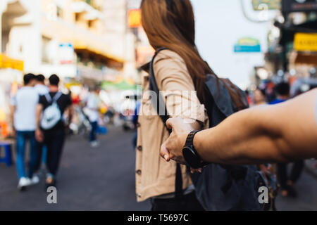 Young girlfriend holding hand of boyfriend. Tourist backpacker couple walking in street market together in Bangkok, Thailand. Follow me concept Stock Photo