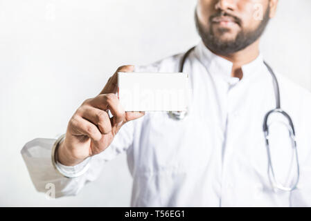 Handsome young doctor in a white coat with a stethoscope holds on the outstretched hand a blank white package box for blister of pills. Mock up. Stock Photo