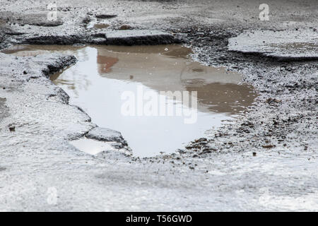 road is paved with cracks in pits and puddles from the snow. Landscape of the winter road in cloudy gray weather. Stock Photo