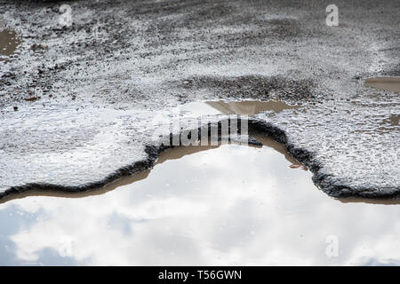 road is paved with cracks in pits and puddles from the snow. Landscape of the winter road in cloudy gray weather. Stock Photo