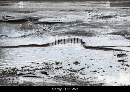 road is paved with cracks in pits and puddles from the snow. Landscape of the winter road in cloudy gray weather. Stock Photo