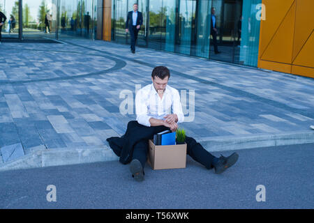 Fired business man sitting frustrated and upset on the street near office building with box of his belongings. He lost work Stock Photo