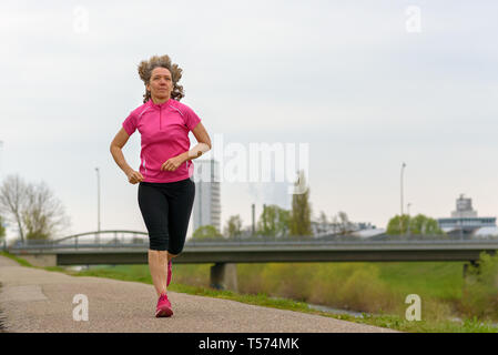Middle age woman in pink training shirt running outdoors down the walkway in park. Full-length low angle front view Stock Photo