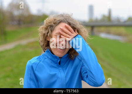 Middle-aged curly woman with curly hair, wearing blue training jacket, smiling, standing and resting with her hand to her forehead after the run. Clos Stock Photo